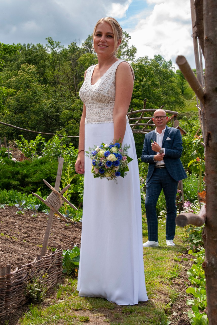 Braut und maßgeschneidertes Brautkleid im Vordergrund, Bräutigam im Hintergrund